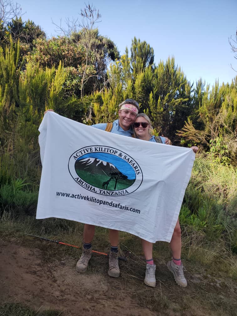 Two climbers holding the Active Kilitop & Safaris flag among giant heather on Mount Kilimanjaro