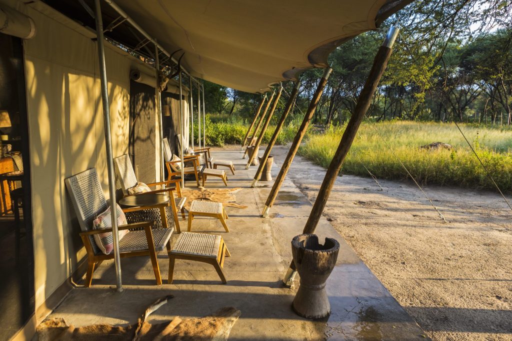 The shaded veranda of a canvas safari tent, with chairs looking out over the grass